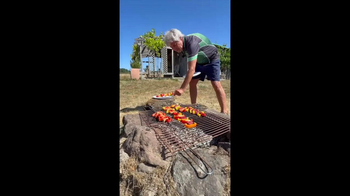Bunte KIVEGAN Spieße auf einem Rost über offenem Feuer, dahinter eine Pergola auf einer Wiese